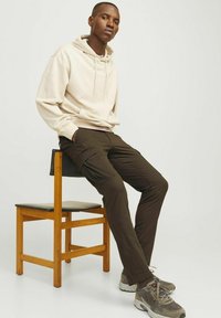 Beige hoodie met trekkoord, gecombineerd met donkere bruine cargobroek met zijzakken. Model zit op een houten stoel.