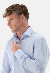 Camicia azzurro chiaro con tessuto testurizzato, maniche lunghe e colletto ripiegato. Dettagli notevoli includono bottoni bianchi e sottili strisce.