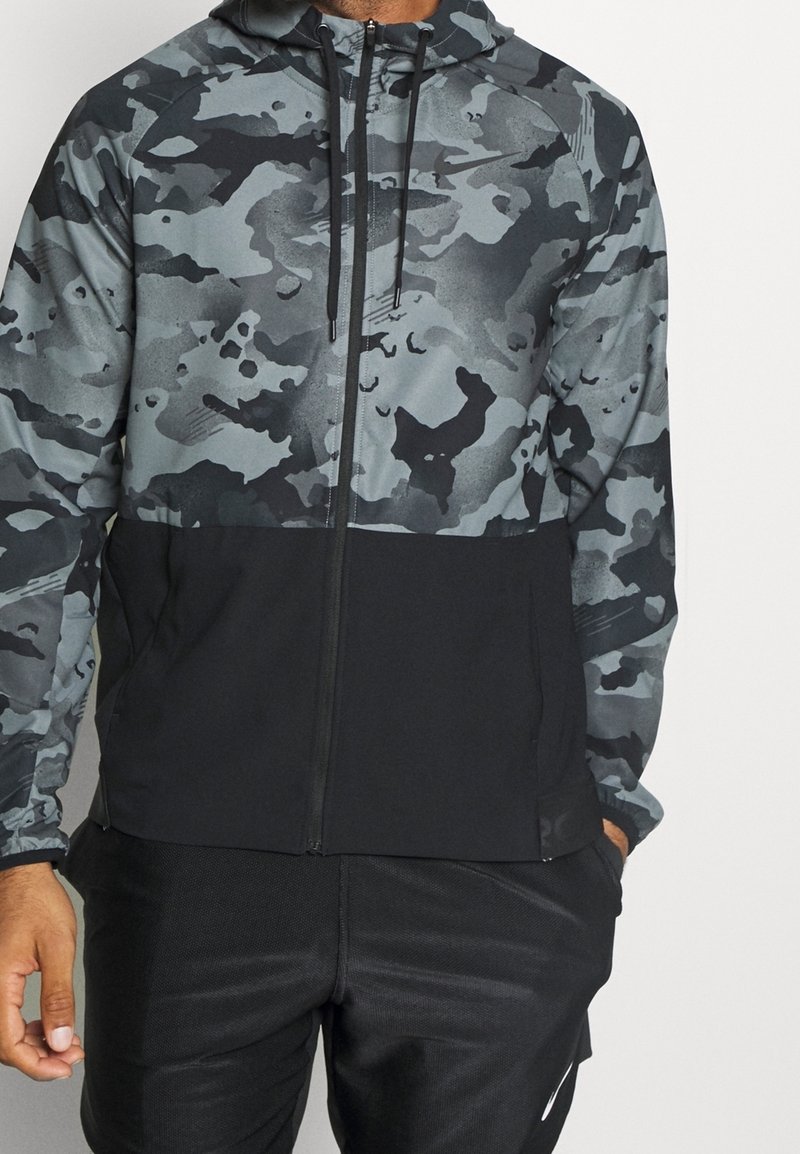 Homme portant un sweat à capuche zippé avec le haut en camouflage gris et noir et le bas uni noir, associé à un pantalon de sport noir.