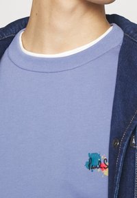 Camicia a maniche corte azzurro chiaro con una finitura testurizzata e colletto interno bianco. Presenta un logo ricamato colorato sul petto. Giacca in denim visibile.