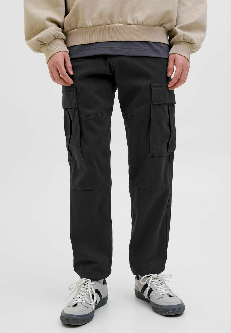 Schwarze Cargo-Hosen mit mehreren seitlichen Taschen, elastischen Bündchen und einem glatten, strapazierfähigen Stoff. Kombiniert mit grauen Sneakers mit schwarzen Akzenten.