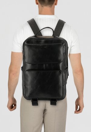 Zaino in pelle nera con spallacci imbottiti, diverse tasche con cerniera e una texture liscia. Presenta un logo prominente e dettagli con fibbie.