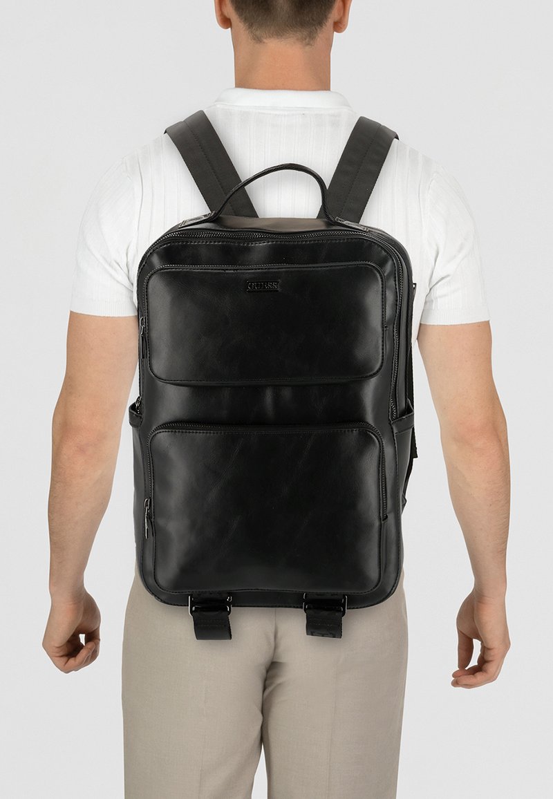 Zaino in pelle nera con spallacci imbottiti, diverse tasche con cerniera e una texture liscia. Presenta un logo prominente e dettagli con fibbie.