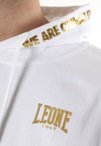Sudadera blanca con detalles dorados. Presenta el logo "LEONE" y el texto "WE ARE COMBAT" en dorado en la capucha. Tela suave con un corte relajado.