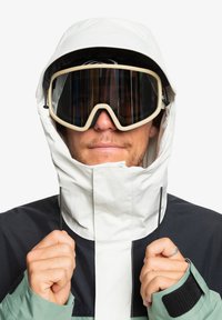 Weiße wasserdichte Jacke mit hohem Kragen, schwarzen Akzenten und verstellbarem Kapuze. Getragen mit großen schützenden Skibrillen. Strukturiertes Gewebe.