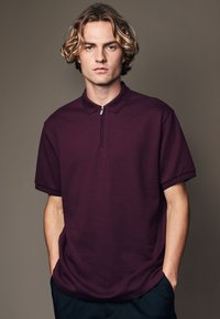 Polo à manches courtes bordeaux avec col zippé, tissu texturé et coupe décontractée. Le modèle se tient les mains dans les poches.