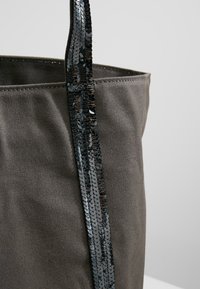 Grijze stoffen tote bag met een textuurafwerking, voorzien van een zwarte paillettenriem die een reflecterende toets toevoegt. Eenvoudig, onopgesmukt ontwerp.