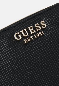 Black textured wallet featuring a gold "GUESS" logo and "EST 1981" detail. Zipper closure with a finely patterned surface.
