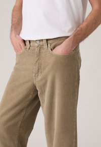 Personne portant un pantalon en velours côtelé beige avec les mains dans les poches avant et un t-shirt blanc, montrée de la taille jusqu'au milieu des cuisses.