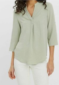 Femme portant un chemisier vert clair à col en V avec des manches trois-quarts et un pantalon blanc, posant devant un fond uni.