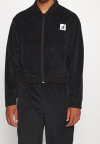 Chaqueta bomber negra con cremallera hecha de un tejido suave y texturizado. Cuenta con un dobladillo y puños acanalados, con un pequeño parche de logo en el lado izquierdo.