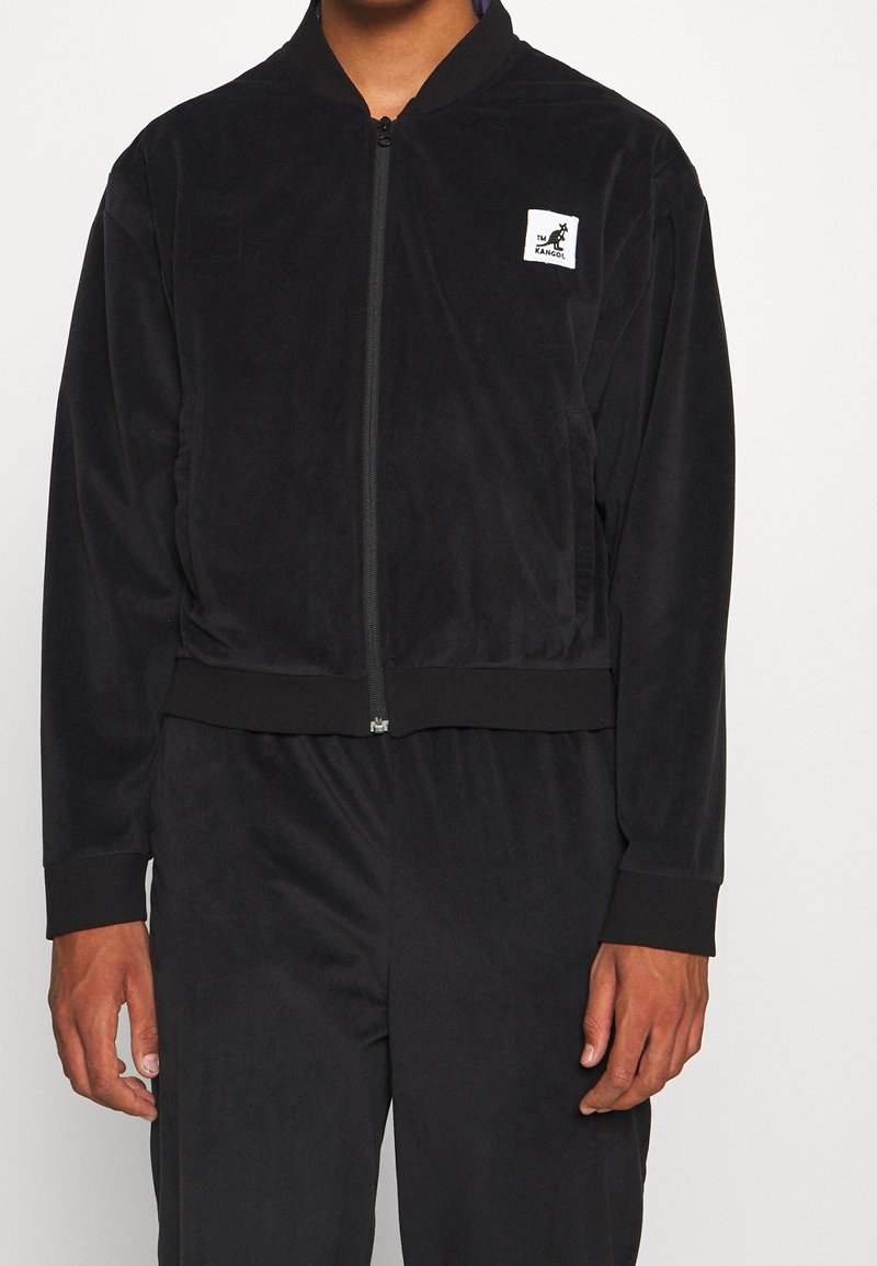 Chaqueta bomber negra con cremallera hecha de un tejido suave y texturizado. Cuenta con un dobladillo y puños acanalados, con un pequeño parche de logo en el lado izquierdo.