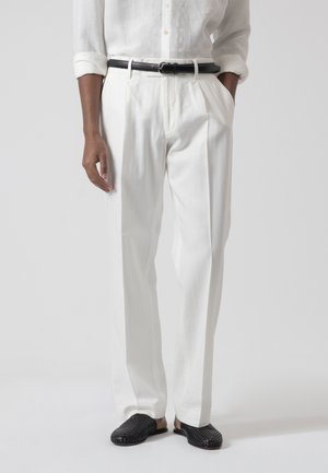 Hombre viste pantalones y camisa de lino blanco con cinturón negro y zapatos slip-on negros trenzados, de pie con una mano en el bolsillo.