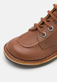 Chaussure en cuir marron avec un bout arrondi, des coutures contrastées, des œillets en métal et une surface texturée. Présente des lacets marron et une semelle en couches.