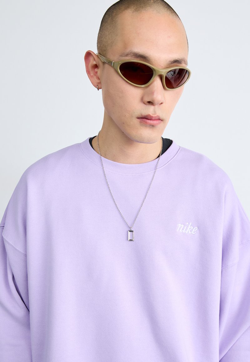 Sweatshirt oversized lavande avec logo "Nike" brodé. Personne porte des lunettes de soleil teintées olive et un collier rectangulaire en argent.