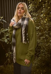 Pull en tricot vert olive avec manches longues, associé à une écharpe à carreaux noir et blanc avec des franges. Arrière-plan de feuillage.