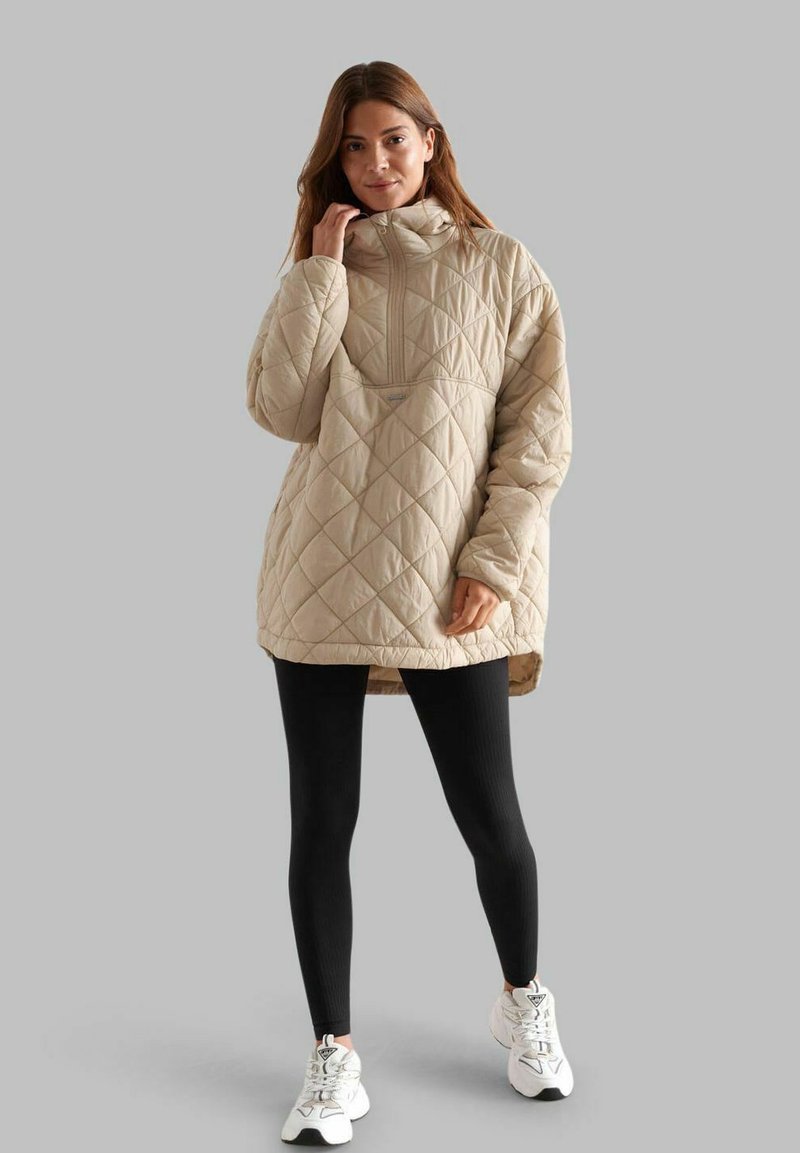 Beige quilted pullover jacket with high collar, front zipper, and side slits, paired with black ribbed leggings and white sneakers.