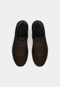 Bruna sneakers med ett material som liknar mocka, svarta läderdetaljer vid hälen och ett platt snörningssystem. Minimalistisk design och slät textur.