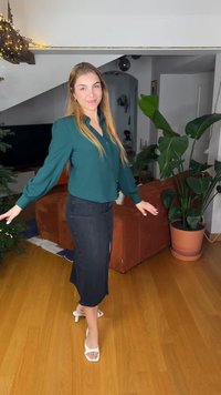 Green button-up blouse with puffed sleeves, black skirt with a slit, and white open-toe heels. Wooden floor, indoor plant, and sofa in the background.