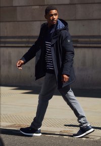 Manteau à capuche bleu marine porté sur une chemise rayée bleu marine et blanche, jeans gris et baskets noires. Marchant sur un trottoir avec un bâtiment en pierre en arrière-plan.