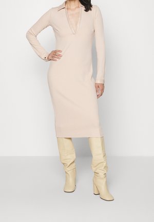 Femme portant une robe midi beige à manches longues avec un décolleté en V profond et des bottes crème à talons montant au genou, debout avec une main sur la hanche.