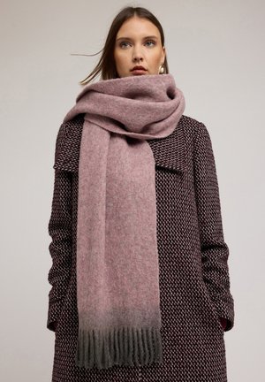 Femme portant une grande écharpe douce rose pâle avec des franges grises sur un manteau bordeaux texturé, debout devant un fond clair uni.