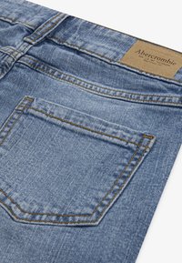 Plave traperice s stražnjim džepom koji ima narančaste šavove i tamniju kožnu oznaku na struku. Tekstura tkanine s izbljedjelim završnim izgledom.