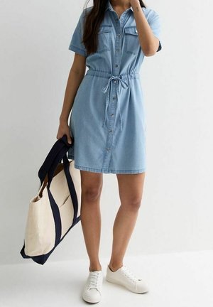 Femme portant une robe chemise en denim bleu clair avec une ceinture à la taille, tenant un sac fourre-tout beige avec des anses bleu marine, et des baskets blanches.