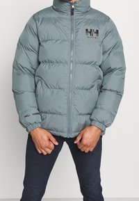 Oliwkowa, pikowana kurtka z wysokim kołnierzem, zapinana na zamek, z elastycznymi mankietami i logo "Helly Hansen" na piersi oraz rękawie.