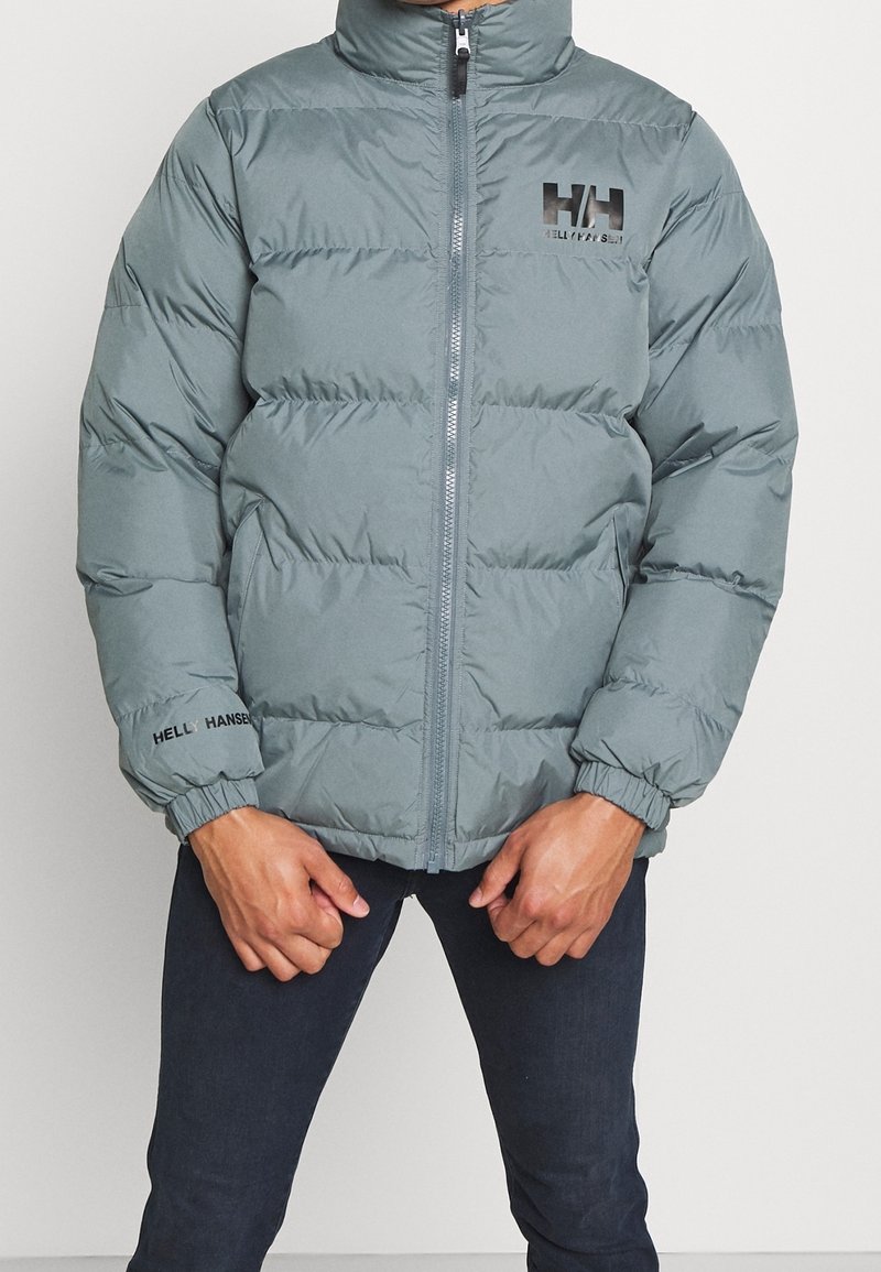 Oliwkowa, pikowana kurtka z wysokim kołnierzem, zapinana na zamek, z elastycznymi mankietami i logo "Helly Hansen" na piersi oraz rękawie.