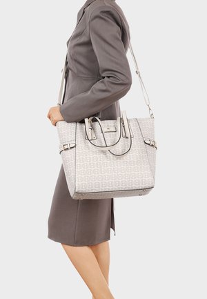 Donna in completo grigio che tiene una grande borsa a spalla grigio chiaro con un motivo a logo ripetuto e tracolla regolabile.