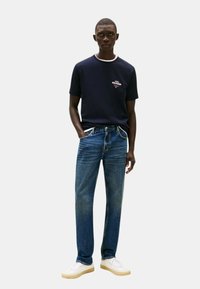 T-shirt crewneck blu navy con accenti bianchi, abbinato a jeans dritti blu e sneaker bianche. Design casual e moderno con dettagli del logo.