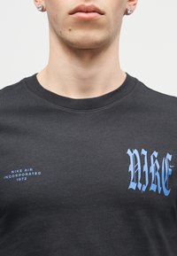 Κοντινό πλάνο ενός ατόμου που φοράει μαύρο μπλουζάκι Nike με μπλε γοτθική γραμματοσειρά "NIKE" και μικρό κείμενο "NIKE AIR INCORPORATED 1972" στο στήθος.