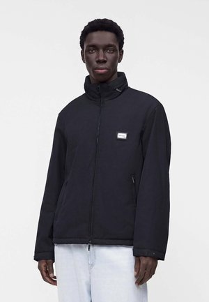 Ung mand iført en sort lynlåstrøje med høj krave og lyseblå jeans, stående mod en ensartet lys baggrund.