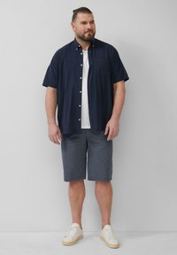 Chemise à manches courtes bleu marine à boutons sur t-shirt blanc ; shorts gris à motifs ; baskets blanches. Coupe ample, tenue décontractée pour l'extérieur.