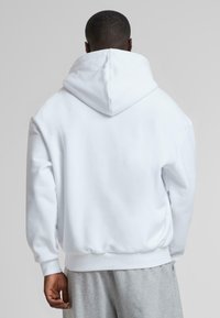 White hoodie made of soft fabric, featuring a large hood, relaxed fit, and ribbed cuffs. Back view shows minimalist design without logos.