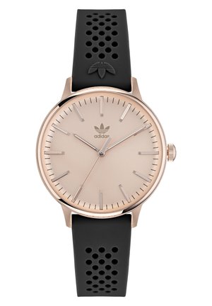 Montre en or rose avec un cadran beige, un boîtier en acier inoxydable, un bracelet en silicone perforé noir et des index d'heure argentés. La marque est visible.