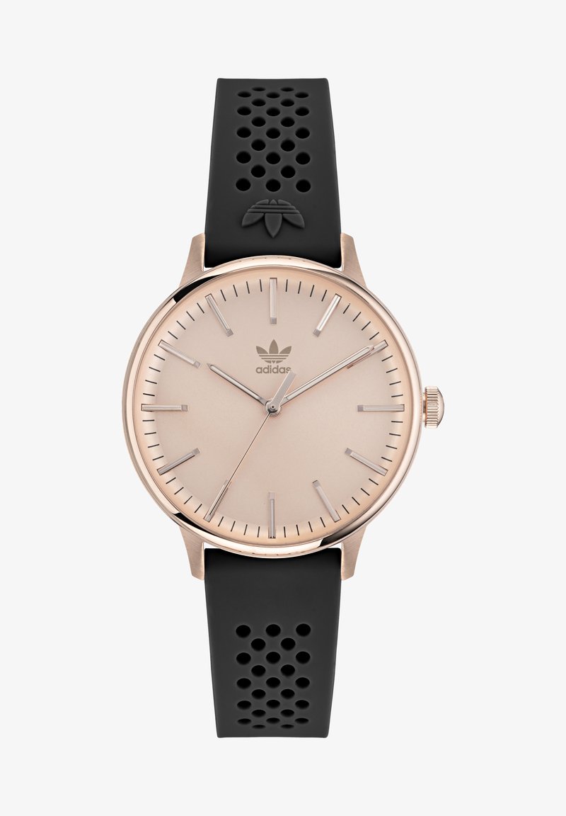 Hodinky v růžově zlaté barvě s béžovým ciferníkem, pouzdrem z nerezové oceli, černým perforovaným silikonovým páskem a stříbrnými hodinovými indexy. Logo značky je viditelné.