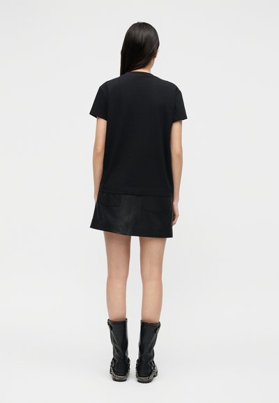 Γυναίκα με μακριά σκούρα μαλλιά, που φοράει μαύρο κοντομάνικο μπλουζάκι, μαύρη δερμάτινη μίνι φούστα και μαύρες μπότες μέχρι τη μέση της γάμπας, στέκεται γυρισμένη με την πλάτη.