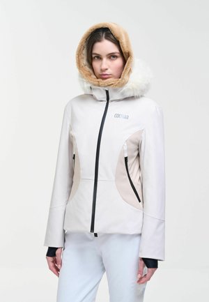 Femme portant une veste d'hiver blanche avec des panneaux beiges et une capuche doublée de fourrure, debout devant un fond clair uni.