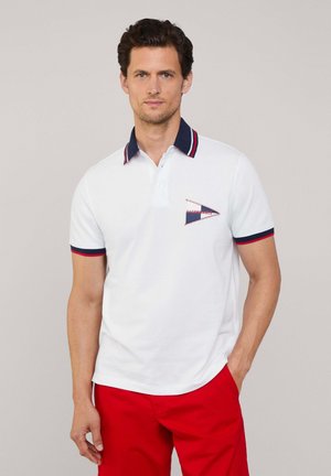 Camisa polo blanca con un cuello azul marino y acentos rojos. Presenta un logo bordado triangular en el lado izquierdo del pecho. Se lleva con pantalones rojos.