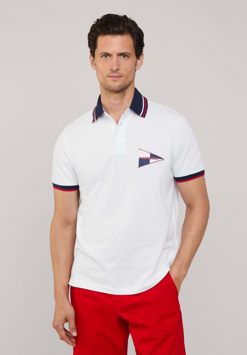 Witte polo met een marineblauwe kraag en rode accenten. Bevat een driehoekig geborduurd logo op de linkerborst. Draagt met rode broek.