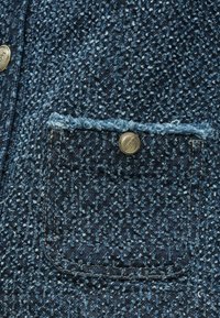 Tessuto di denim blu con un aspetto strutturato, caratterizzato da una tasca frontale, bordi sfrangiati e una chiusura con bottone a pressione metallico.
