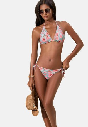 Nainen, jolla on päällään sininen ja pinkki kukkakuvioinen bikini sivusolmuille, pitämässä ruskeaa punottua hattua, ja jolla on suuret neliskulmaiset aurinkolasit sekä rannekoru.