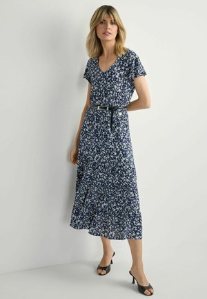 Kobieta ubrana w granatową kwiecistą sukienkę midi z czarnym paskiem i czarnymi sandałami na obcasie z otwartymi palcami, stojąca na tle jasnoszarej, gładkiej ściany.