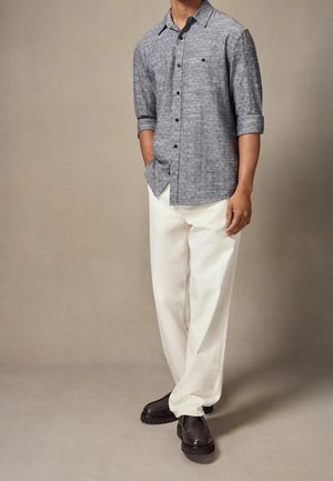 Pánská košile na knoflíky ze strukturované šedé látky s ohrnutými rukávy, kombinovaná s volnými bílými kalhotami a černými robustními botami.