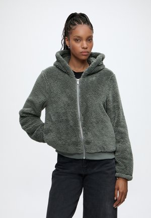 Femme portant une veste polaire grise zippée à capuche et un pantalon noir, debout devant un fond blanc uni.