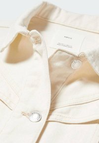 Cremefarbene Denimjacke mit strukturiertem Kragen, silbernen Druckknöpfen und sichtbaren Nähdetails an der Brusttasche und den Nähten.