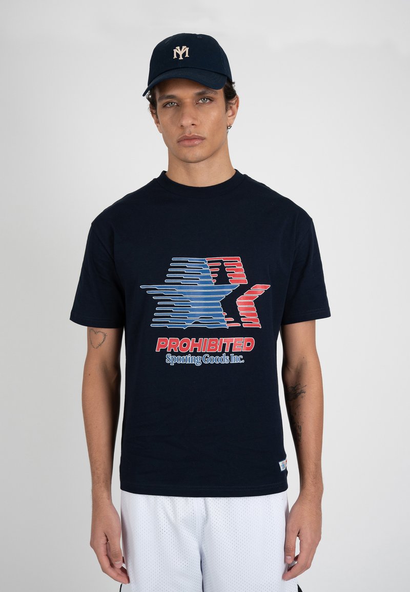 Joven con gorra azul marino y camiseta azul marino con gráfico de estrella en rojo, blanco y azul y texto "PROHIBITED Sporting Goods Inc."