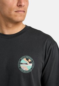 Black cotton t-shirt featuring a circular logo in pastel colors, depicting a landscape with waves, sun, and "Billabong Adventure Division" text.
